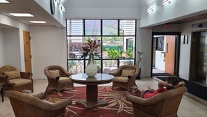 A lobby with wicker furniture, a central round table, and a large window with a view of greenery outside.