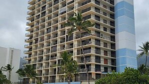 A multi-story hotel with balconies, located near a sandy beach with palm trees.