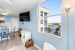 Habitación con televisor montado en la pared, ventana con vista al río y edificios, ventilador de techo y comedor con sillas y mesa.