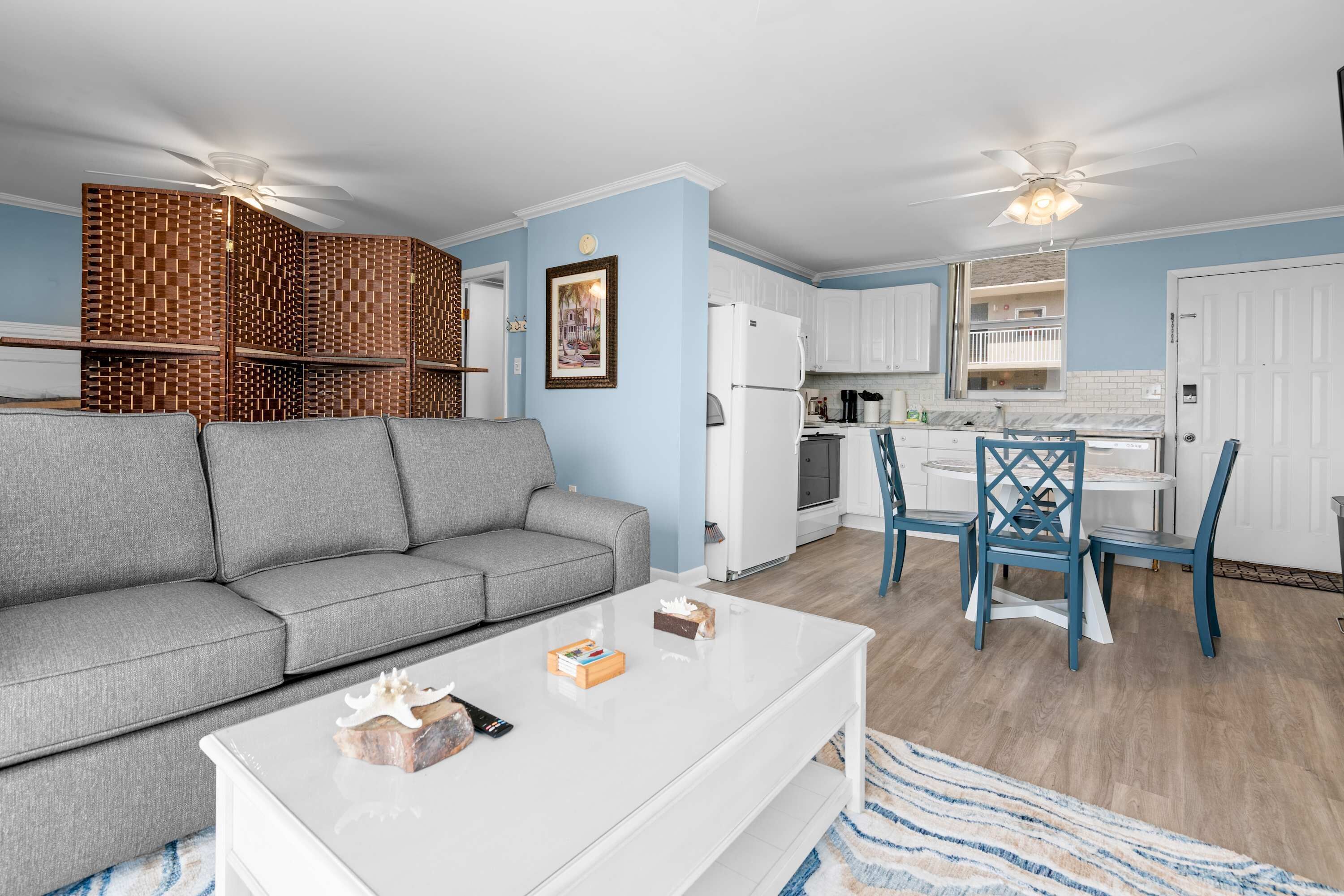 Una sala de estar con un sofá gris, una mesa de centro blanca y una zona de cocina con armarios blancos y sillas azules.