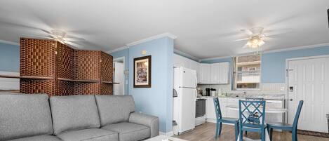 Una sala de estar con un sofá gris, una mesa de centro blanca y una zona de cocina con armarios blancos y sillas azules.