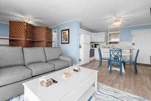 A living room with a grey sofa, a white coffee table, and a kitchen area with white cabinets and blue chairs.