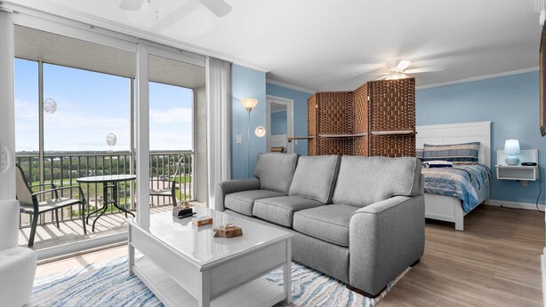 Una camera moderna con un divano grigio, un tavolino bianco, un letto con copriletto a righe blu e bianche, un piccolo tavolo con una lampada e un balcone con vista.