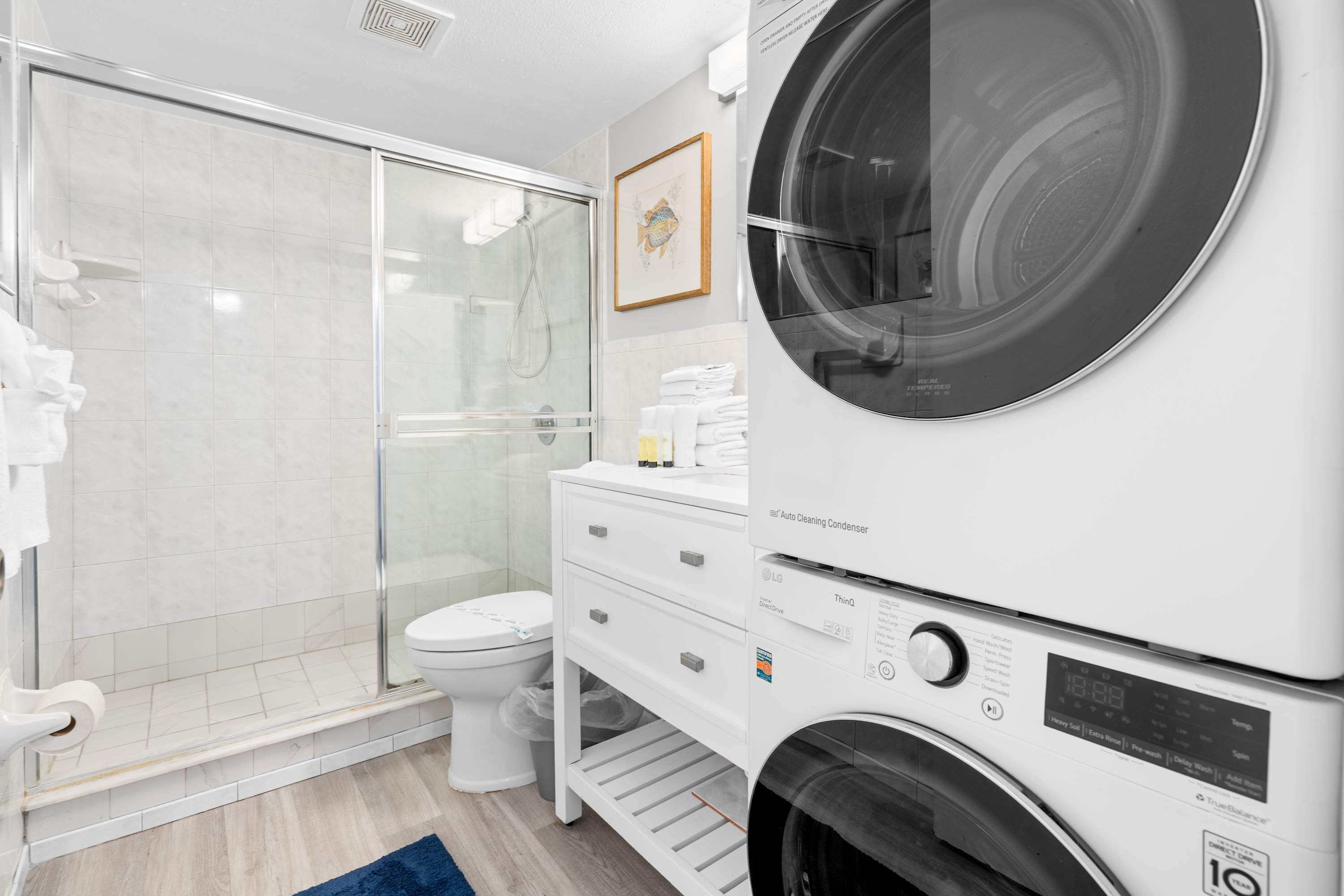 A modern bathroom with a glass-enclosed shower, a toilet, a stacked washer and dryer, and a blue rug.