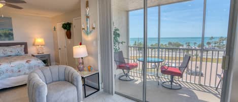 Hotelkamer met een bed, een stoel, een tafel, een balkon met uitzicht op zee en een plafondventilator.
