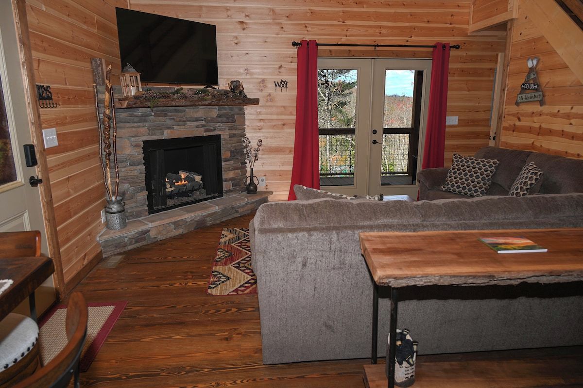 Un acogedor interior de cabaña con chimenea de piedra, un sofá gris y una mesa de madera.