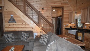Interior acolhedor de uma cabana, com escada de madeira, sofá cinza e cozinha.