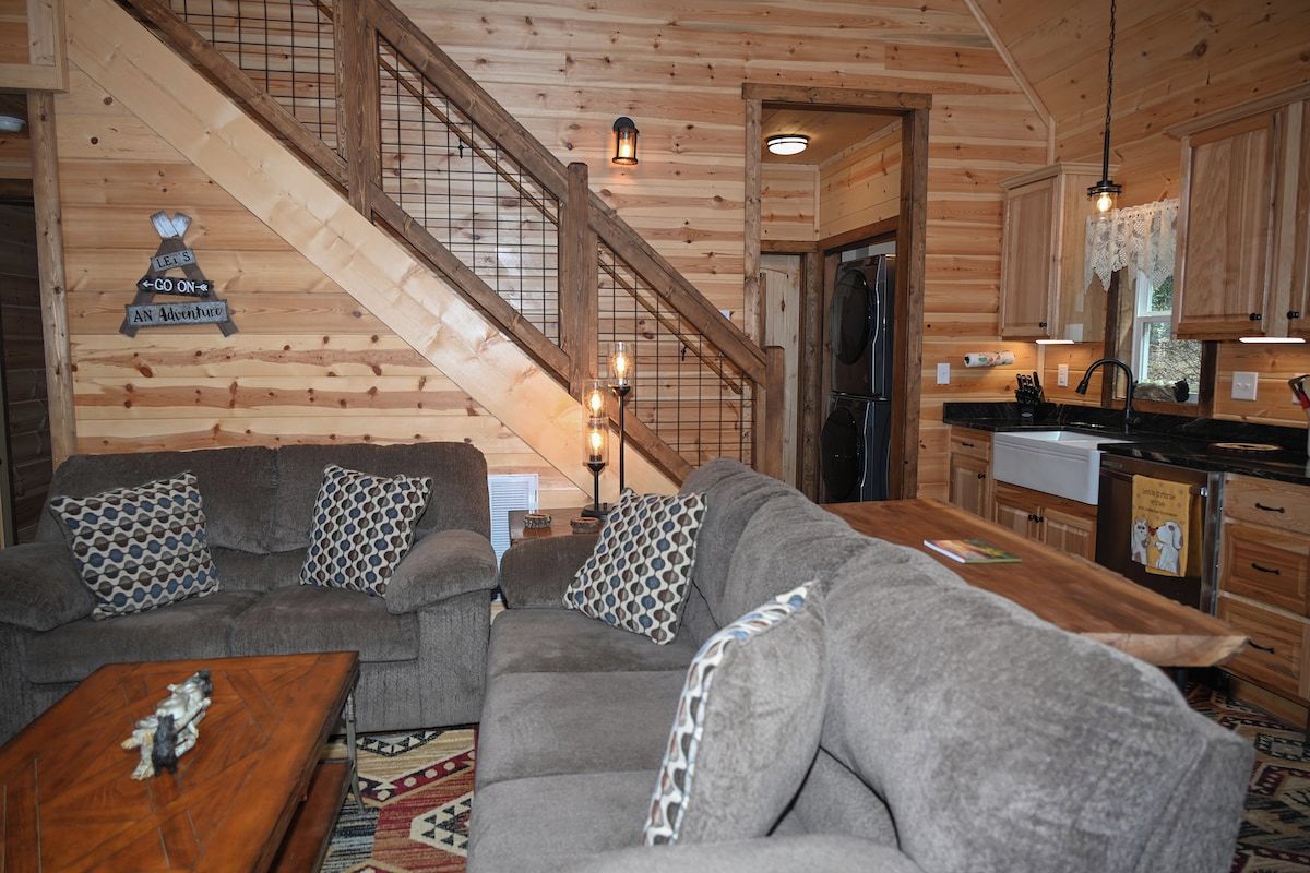 A wooden interior with a staircase, a grey sofa set, a wooden dining table, and a kitchen area.