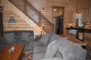 A wooden interior with a staircase, a grey sofa set, a wooden dining table, and a kitchen area.