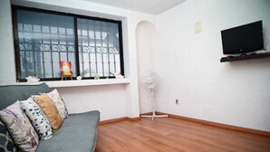 Een woonkamer met een grijze bank, een ventilator en een aan de muur bevestigde televisie.