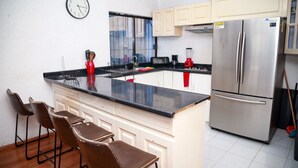 Een keuken met een roestvrijstalen koelkast, een magnetron, een spoelbak en een klok aan de muur. Drie barkrukken en een raam met jaloezieën.