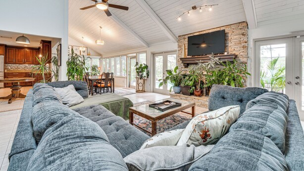 A spacious living room with a large gray sectional sofa, a coffee table, and a fireplace with a TV above it. There are multiple windows providing natural light and a ceiling fan. - 10 Mi to Disney World, Lakeside Orlando Getaway (Orlando)