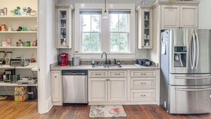 Una cocina con armarios blancos, un refrigerador de acero inoxidable, un lavavajillas y un ventanal con vista a árboles.