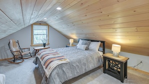 A bedroom with a sloped ceiling, a bed with a patterned bedspread, a rocking chair, a small table with a lamp, and a window with blinds. - Steps to Lake Access! Eastbrook Family Cabin (Eastbrook)