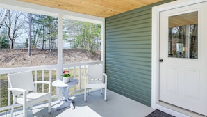 A covered porch with white wicker chairs and a small table, a green door, and a view of trees and a yard. - Steps to Lake Access! Eastbrook Family Cabin (Eastbrook)