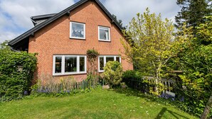 A two-story brick house with a sloped roof, two windows on the upper floor, and a well-maintained garden with grass, shrubs, and a tree. - Apartment 'Ferienwohnung Flensburg Mürwik' with Shared Garden, Balcony and Wi-Fi (Flensburg)