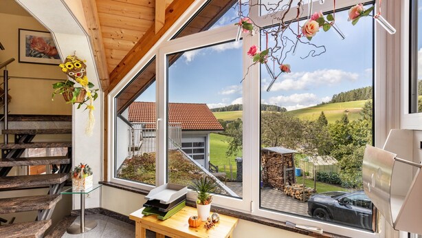 A modern interior with a wooden staircase, a glass table with a plant, and a large window offering a view of a house and landscape. - Holiday Apartment "Dachspitz - Junges Wohnen" with Shared Garden & Wi-Fi (Unterkirnach)