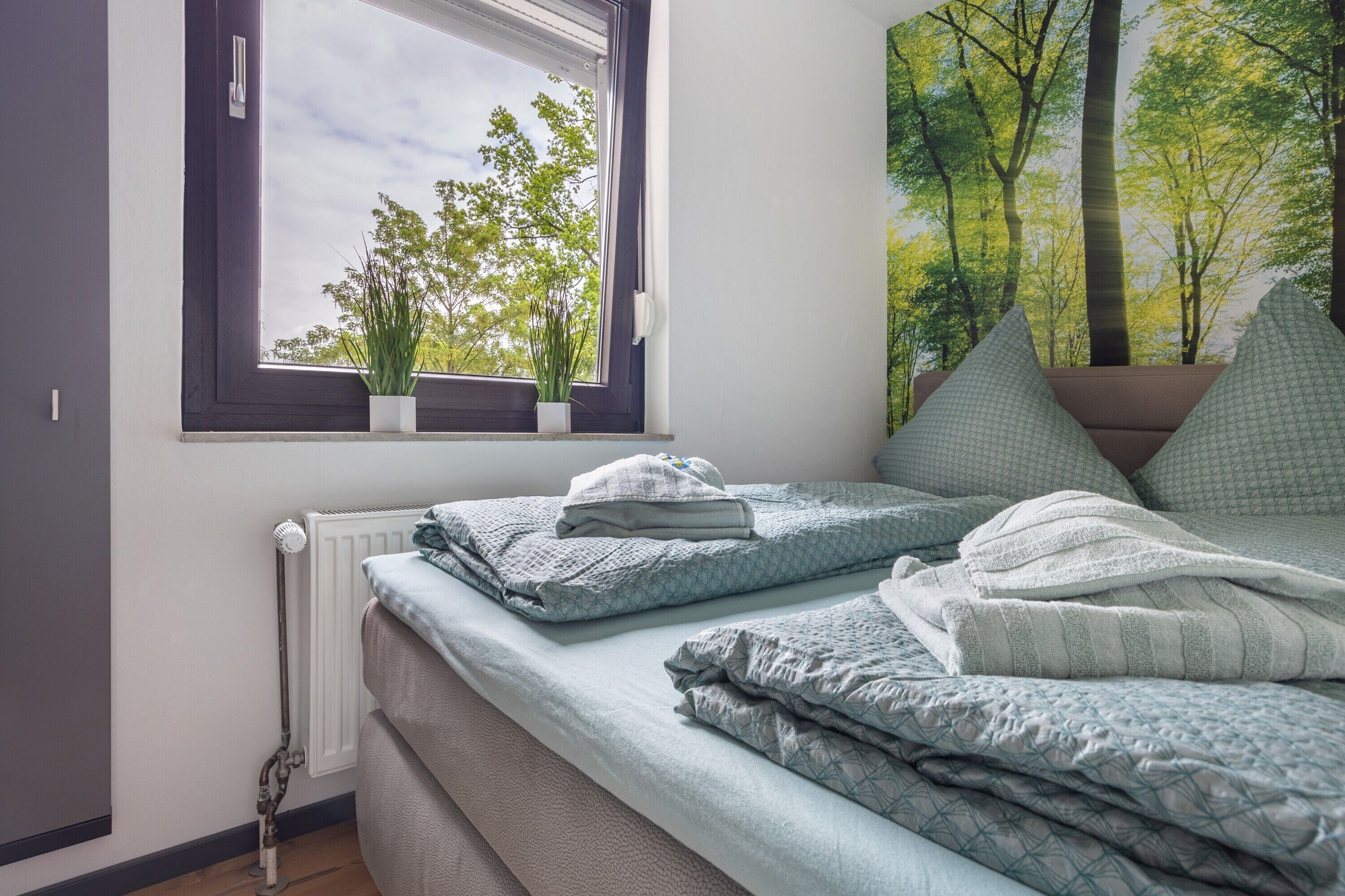 Une chambre à coucher avec un lit, un radiateur, une fenêtre ornée de plantes et un mur décoré sur le thème de la forêt.