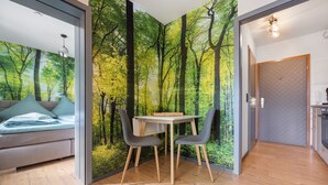 Une pièce moderne avec un plancher en bois, une table à manger avec des chaises, un canapé avec des coussins et une grande fresque représentant une forêt sur le mur.