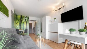 Un salon moderne avec un canapé gris, une table à manger agrémentée de deux plantes en pot, un coin cuisine avec un téléviseur et une fresque représentant une forêt sur le mur.