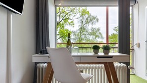 Une chambre moderne avec un bureau blanc, une chaise blanche, un radiateur et une fenêtre donnant sur des arbres.