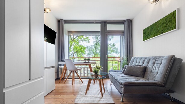 Un salon moderne comprenant un canapé, une table à manger avec des chaises, une petite table avec une plante et une grande fenêtre donnant sur des arbres.