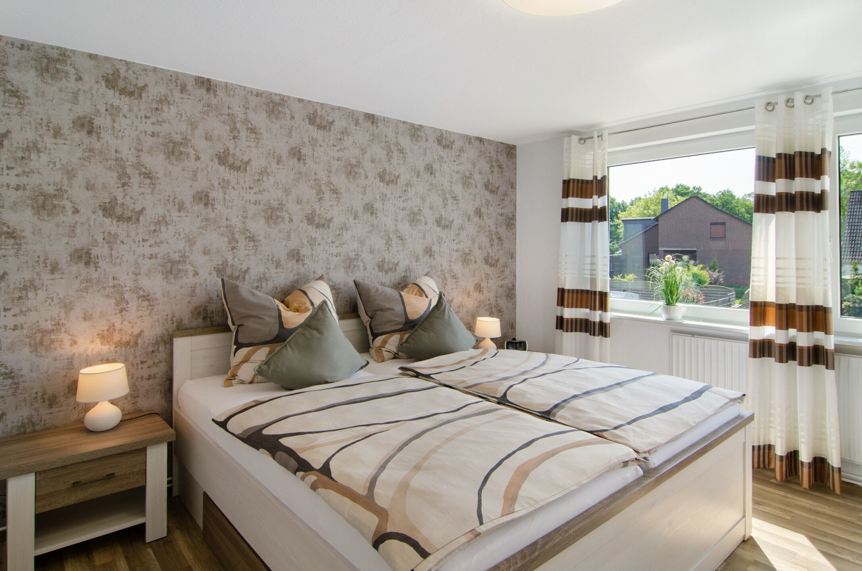 Ein Schlafzimmer mit Bett, Nachttischen, einer Lampe, einem Fenster mit Vorhängen und Blick auf ein Gebäude.
