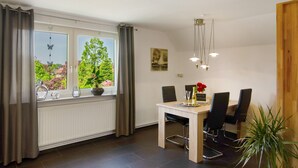 Ein Essbereich mit Tisch, Stühlen, einem Fenster mit Vorhängen, einem Heizkörper und einer Pflanze.