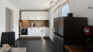 Eine kompakte Küche mit weißen Schränken, einem Edelstahlkühlschrank, einer Kaffeemaschine und einem Fenster mit Jalousien.