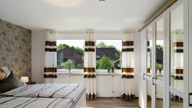 Ein Schlafzimmer mit einem Bett, einem Fenster mit Vorhängen, einer Heizung und Blick ins Freie.