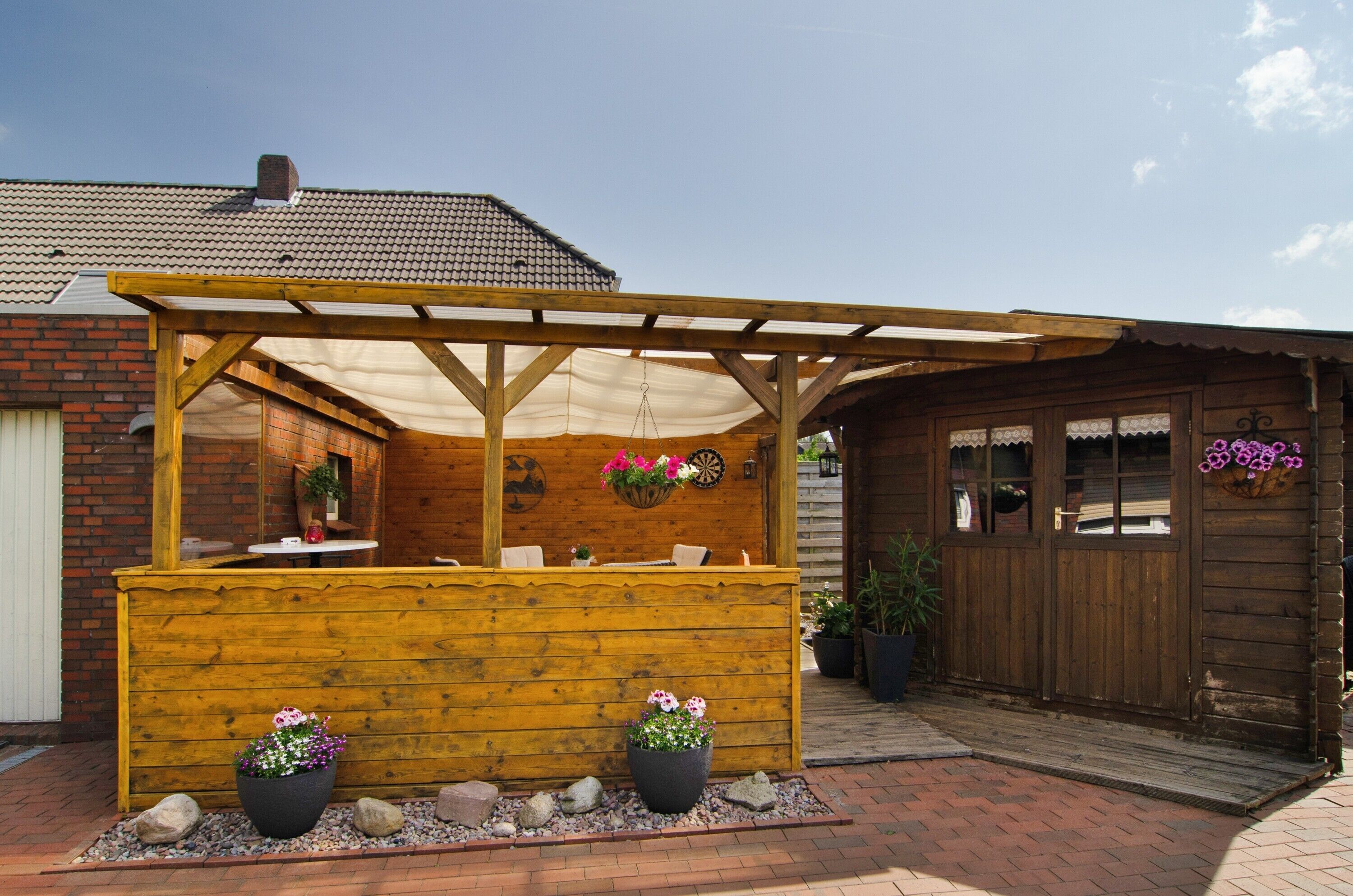 A wooden outdoor seating area with a pergola, potted plants, and a brick building in the background.