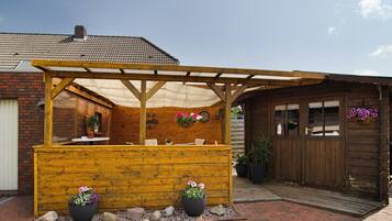 A wooden outdoor seating area with a pergola, potted plants, and a brick building in the background.