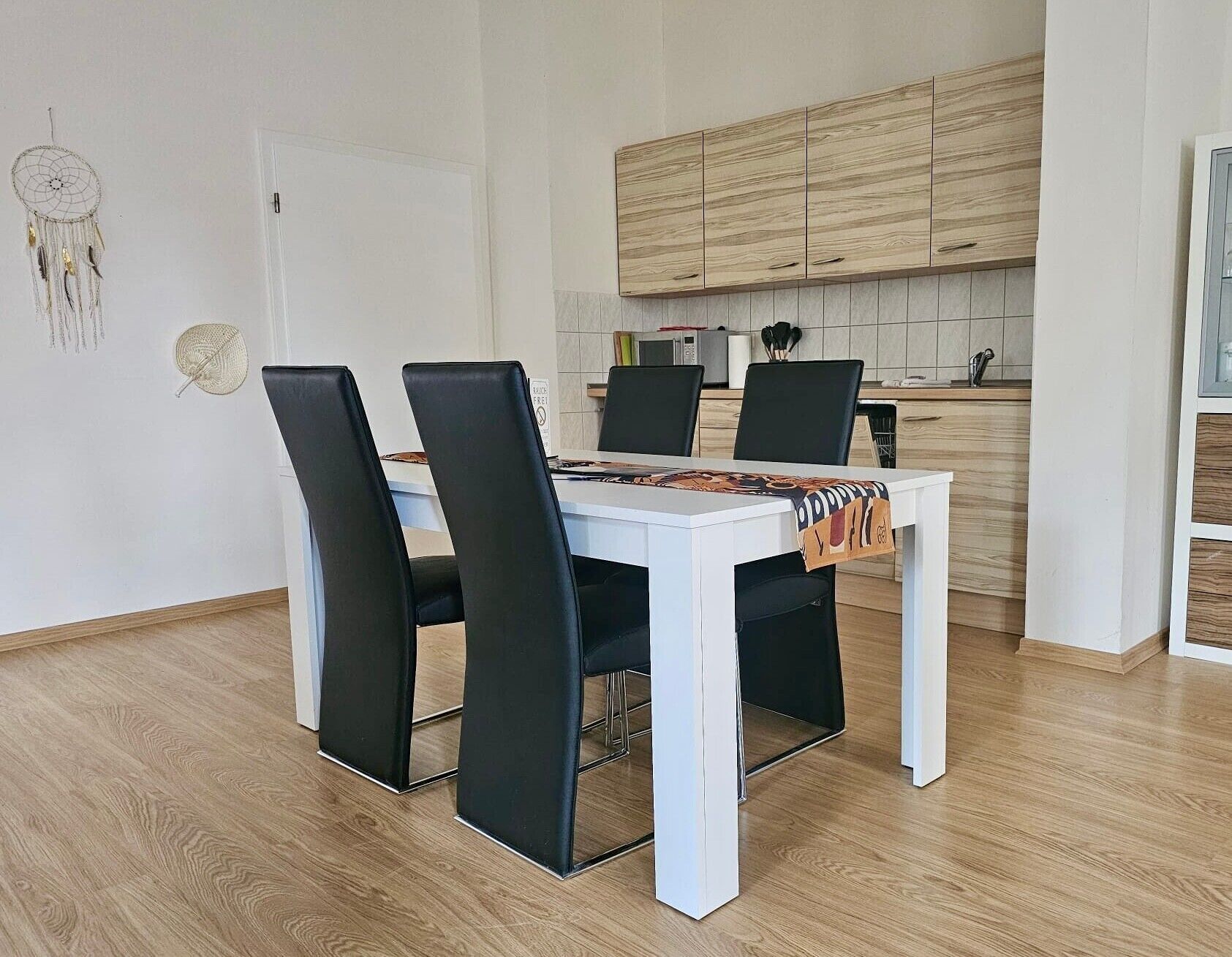 Uma área de refeições com mesa branca e cadeiras pretas, uma cozinha com armários de madeira e um sonho na parede.