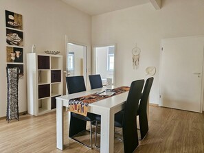 A dining area with a white table, black chairs, and a coffee-themed table runner. There are shelves with decorative items and a dreamcatcher on the wall.