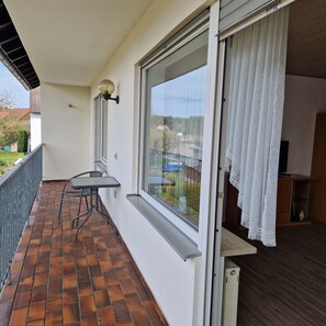 A balcony with a table and chairs, a window with curtains, and a view of a garden and buildings.