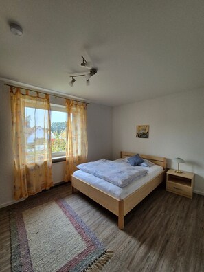 Un dormitorio con cama, mesita de noche, lámpara, ventana con cortinas, luz en el techo y un cuadro en la pared.