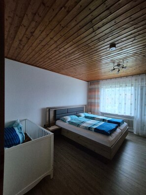 A bedroom with a wooden ceiling, a bed with blue bedding, a nightstand, a crib, and a window with curtains.