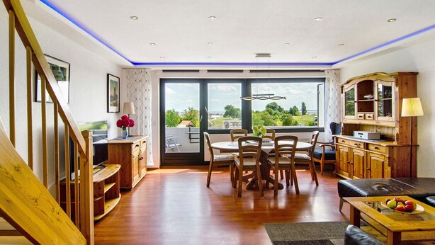 A spacious living area with a dining table, chairs, a sofa, and a kitchen area with wooden cabinets and a view through large windows. - Apartment 'Ferienwohnung Dünenblick' with Sea View, Balcony and Wi-Fi (Wangerland)