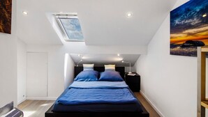 A bedroom with a skylight, a bed with blue bedding, a nightstand, and a wall-mounted painting. - Apartment 'Ferienwohnung Dünenblick' with Sea View, Balcony and Wi-Fi (Wangerland)