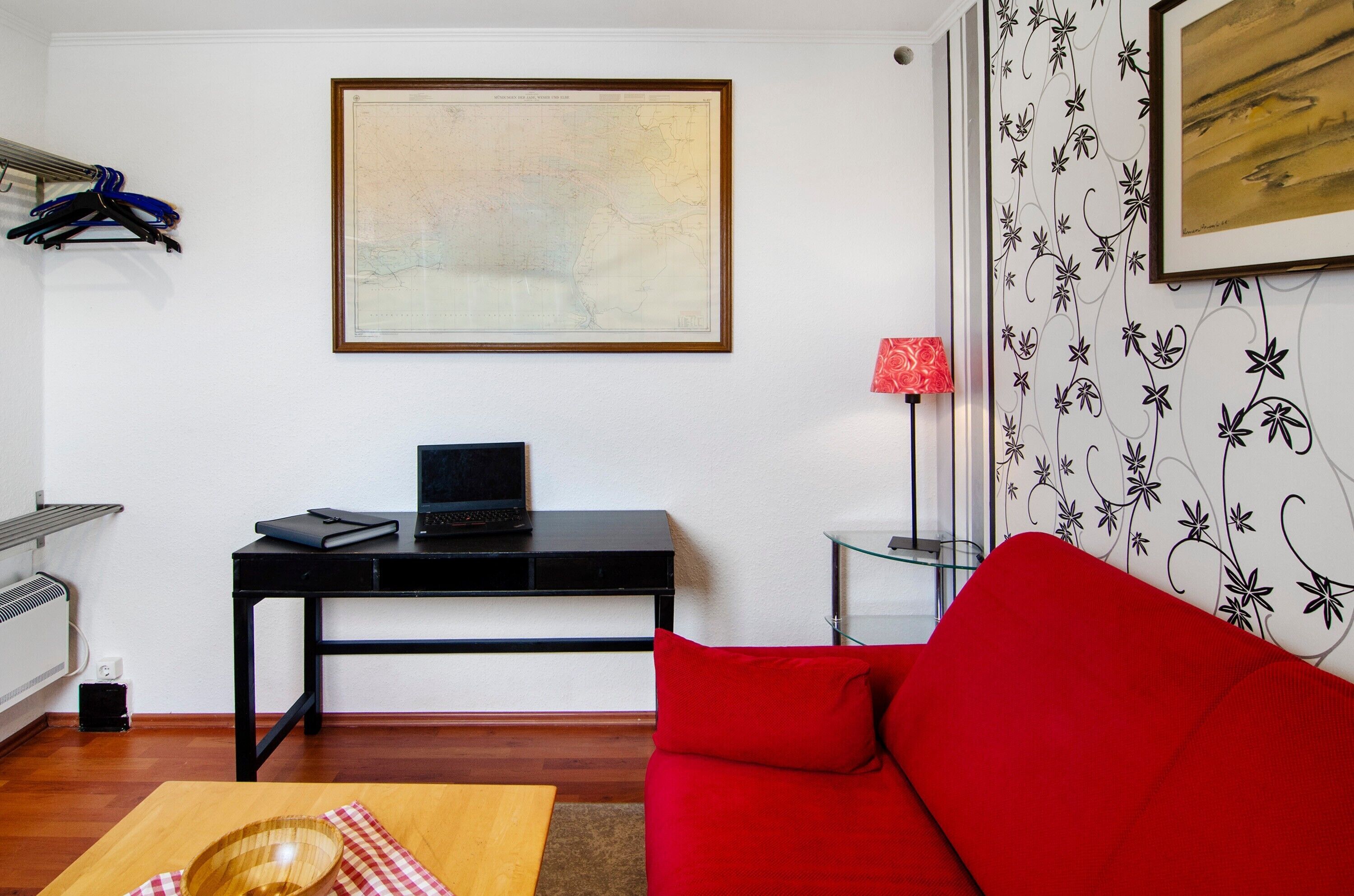 Una habitación con un escritorio negro, un sofá rojo, un mapa enmarcado, una lámpara roja y una pared con estampado floral.