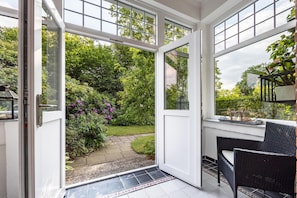 Une véranda dotée d’une porte vitrée donnant sur un jardin, d’un fauteuil en osier et d’une petite table agrémentée d’objets décoratifs.