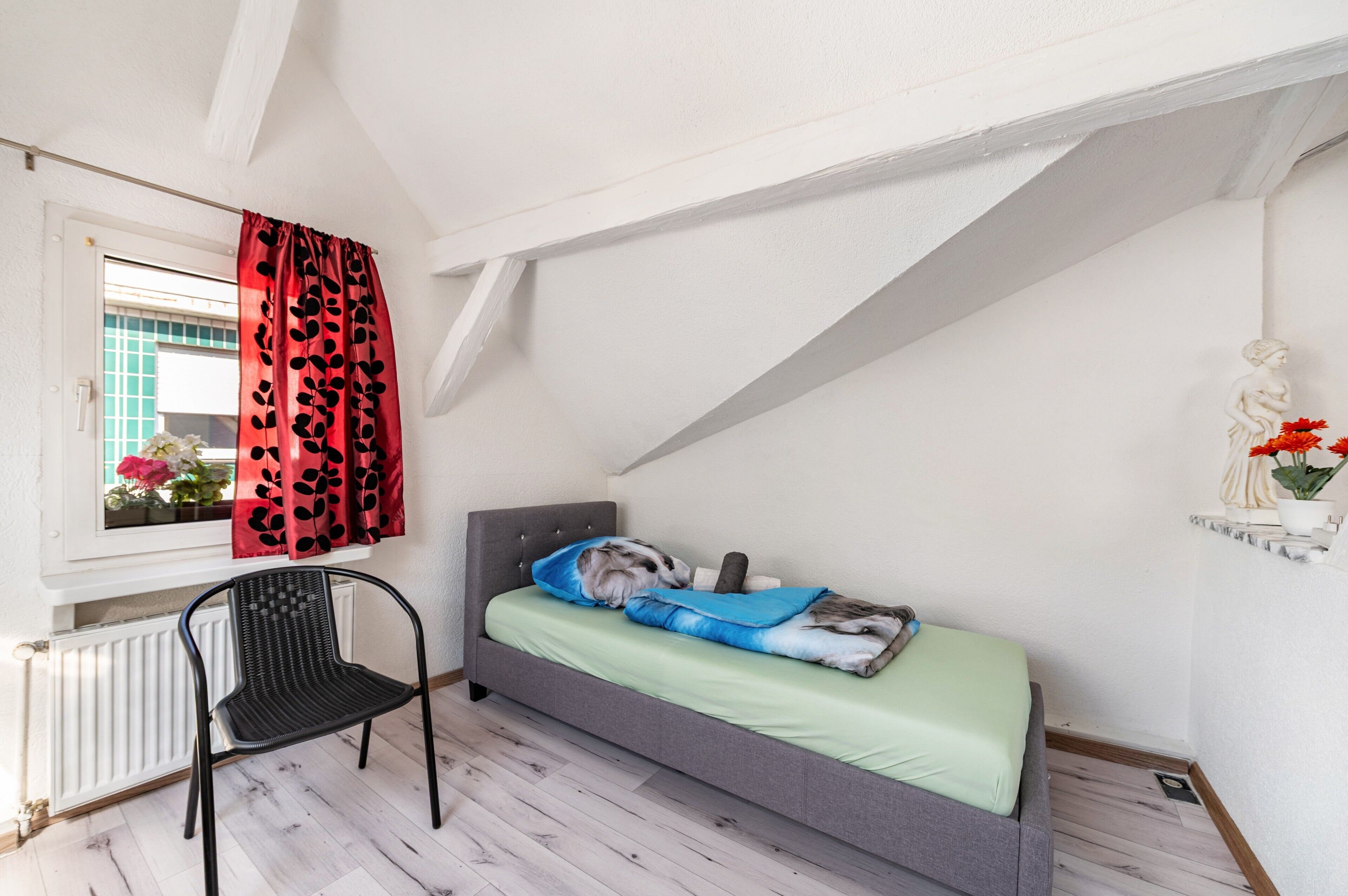 Ein kleines Schlafzimmer mit einem Bett, einem Sessel, einem Fenster mit Vorhängen und einer Heizung.