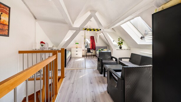 Une pièce mansardée spacieuse avec un escalier en bois, un coin repas avec des meubles en osier, un lit et une grande fenêtre.