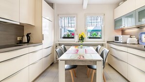 Une cuisine moderne avec des meubles blancs, une table à manger avec des chaises, une bouilloire, un four à micro-ondes et une fenêtre avec des rideaux.