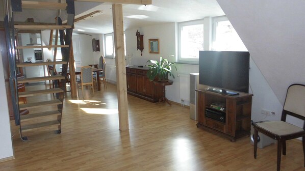 A spacious living area with a wooden staircase, a dining table, chairs, a television, and a potted plant.
