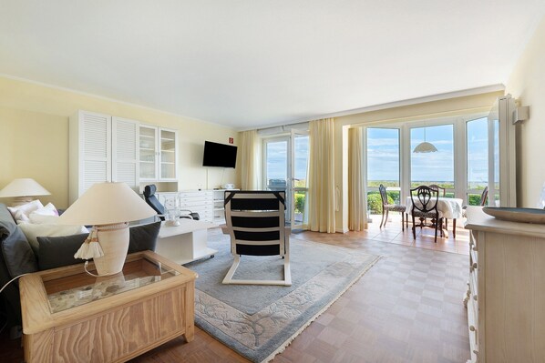 A living room with a sofa, a coffee table, a TV, a dining area with chairs, and a large window with a view.