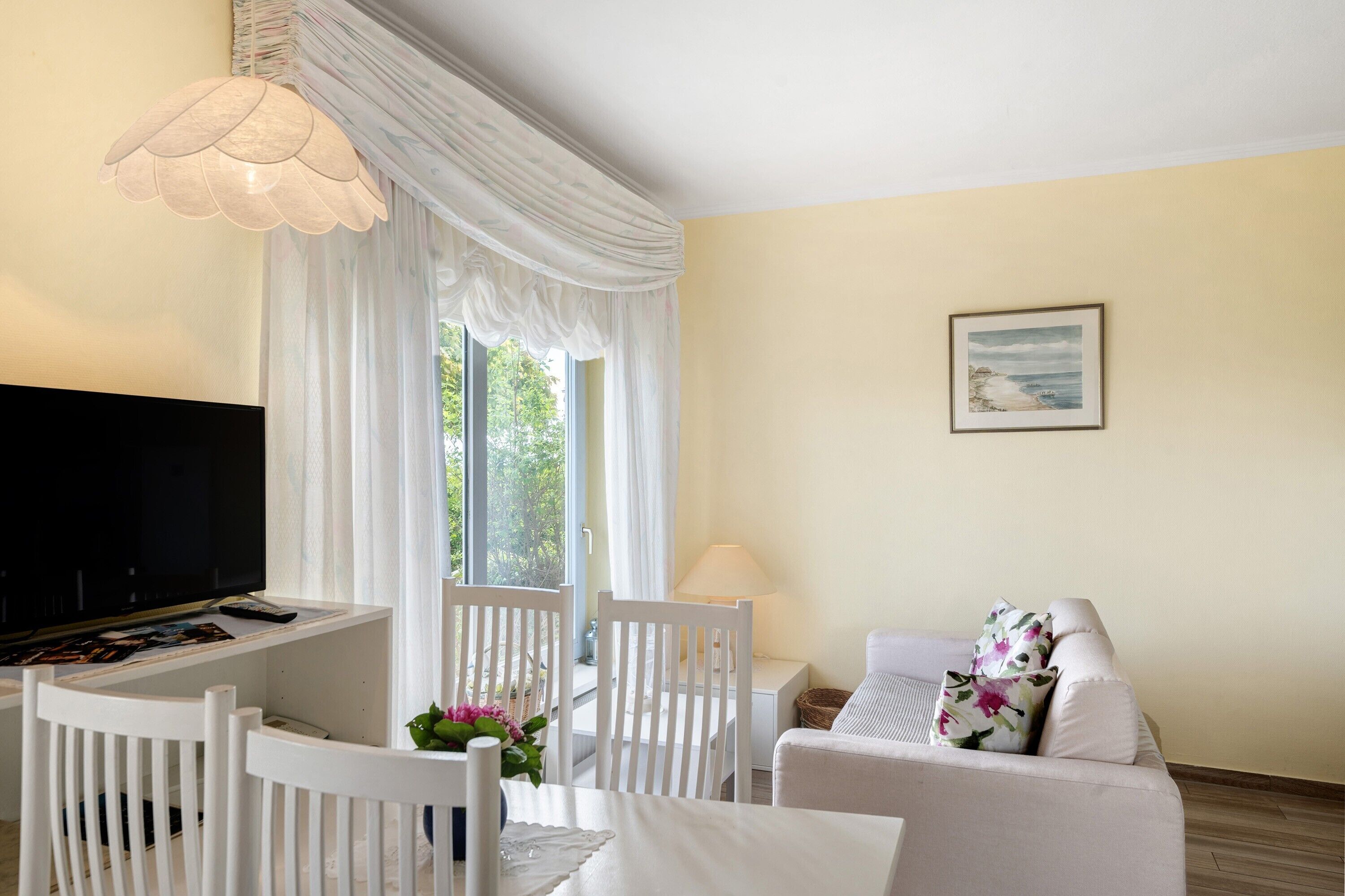 Ein Wohnzimmer mit Fernseher, weißen Möbeln, einer Lampe und einem Fenster mit Vorhängen.