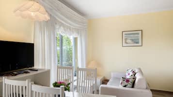 Ein Wohnzimmer mit Fernseher, weißen Möbeln, einer Lampe und einem Fenster mit Vorhängen.