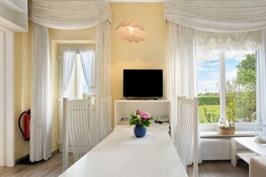 A room with a TV on a stand, a white table with a vase of flowers, and a window with a view of greenery. - Apartment 'Wohnung 2' with Private Terrace, Shared Garden and Wi-Fi (Hohwacht (Ostsee))