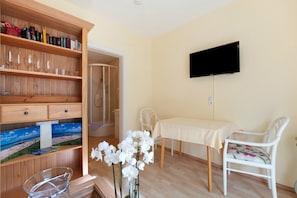 Une pièce avec une bibliothèque en bois, une table recouverte d’une nappe blanche, une chaise blanche, un téléviseur fixé au mur et un espace douche visible à travers une porte ouverte.